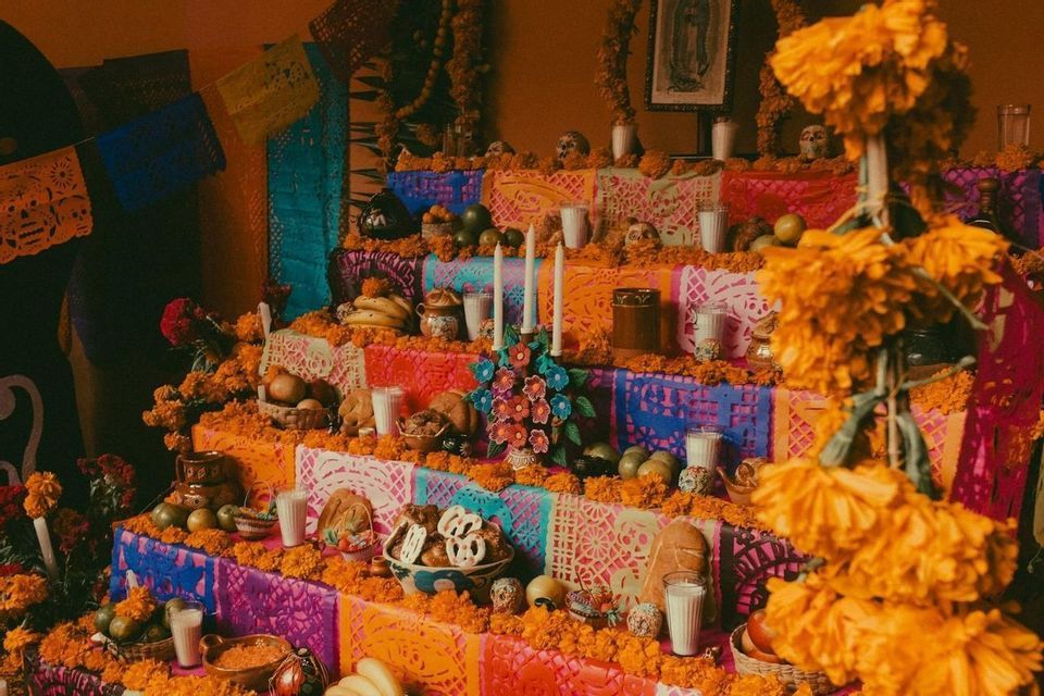 A multi-tiered Day of the Dead altar decorated with orange marigolds, skulls, candles, food, and colorful papel picado.