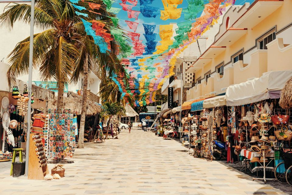 Una strada lastricata fiancheggiata da negozi e palme è decorata con bandiere di carta colorate appese in alto.