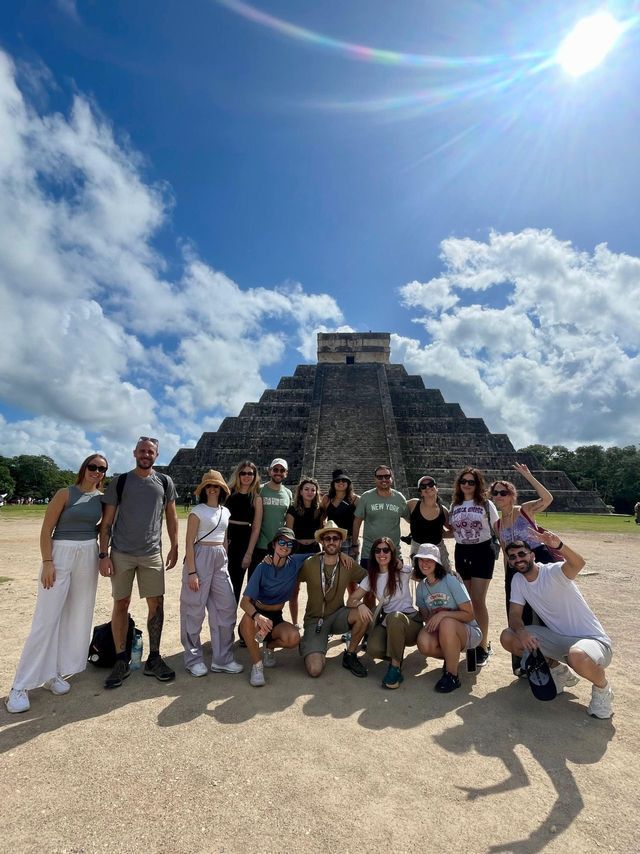 Un viaggio di gruppo WeRoad in posa per una foto davanti a una grande piramide di pietra sotto un cielo soleggiato con nuvole sparse.
