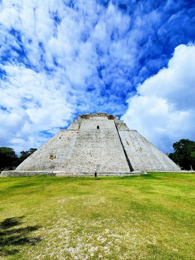 Vista dal basso di una grande piramide di pietra con una persona alla base, su un campo erboso e un cielo blu con nuvole.