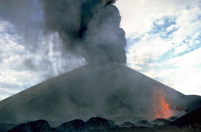 Ein aktiver Vulkan bricht aus und schleudert eine große Säule dunklen Rauchs und Asche in einen bewölkten Himmel, während geschmolzene Lava seine Hänge hinabfließt.