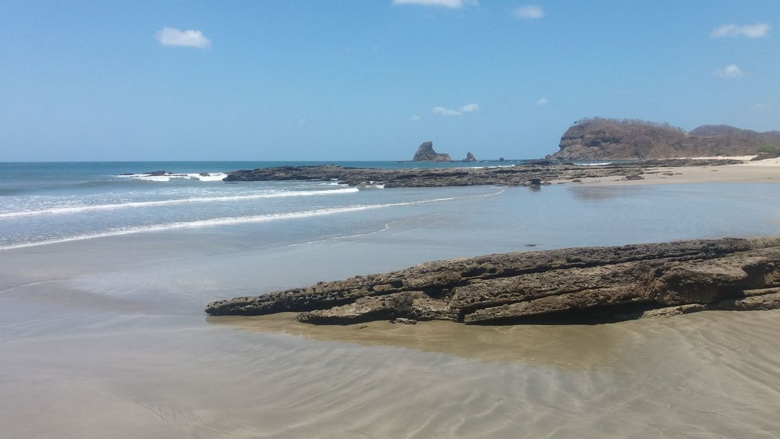 Ein Sandstrand mit großen, dunklen Felsformationen am Ufer und im Meer unter einem klaren blauen Himmel.