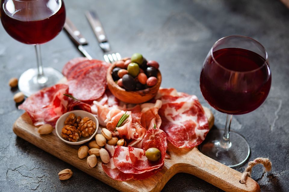 Se sirve una tabla de charcutería de madera con embutidos, aceitunas y frutos secos, acompañada de dos copas de vino tinto.