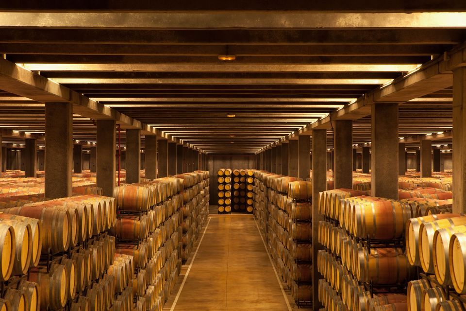 Barriles de madera apilados en largas filas simétricas dentro de una gran bodega, iluminados por cálidas luces cenitales.