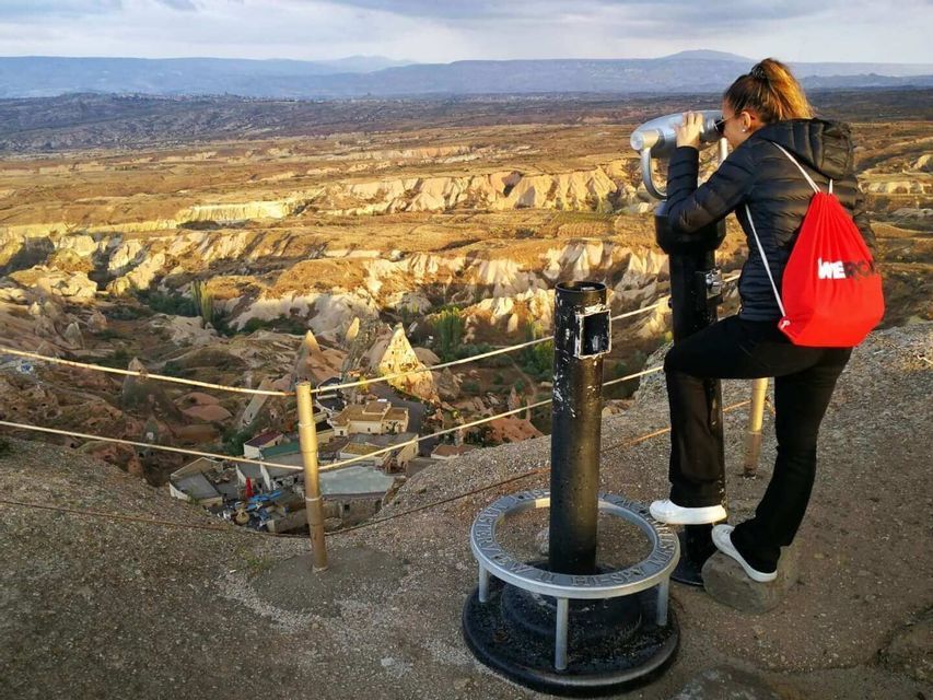 A woman with a red WeRoad backpack looks through a viewfinder, overlooking a vast rocky valley from a high viewpoint.