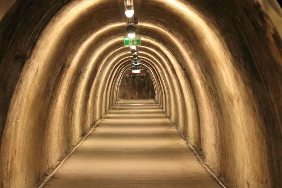 A long, arched underground tunnel with a concrete floor, illuminated by a row of ceiling lights creating a receding perspective.