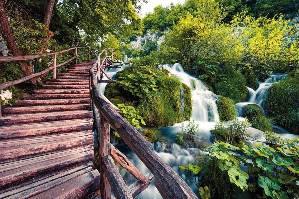 Una scala rustica in legno con ringhiera si snoda accanto a una cascata impetuosa in una foresta verde lussureggiante.