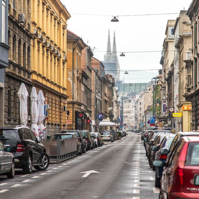Eine lange Stadtstraße mit geparkten Autos und historischen Gebäuden, im Hintergrund zwei Domtürme unter bedecktem Himmel.
