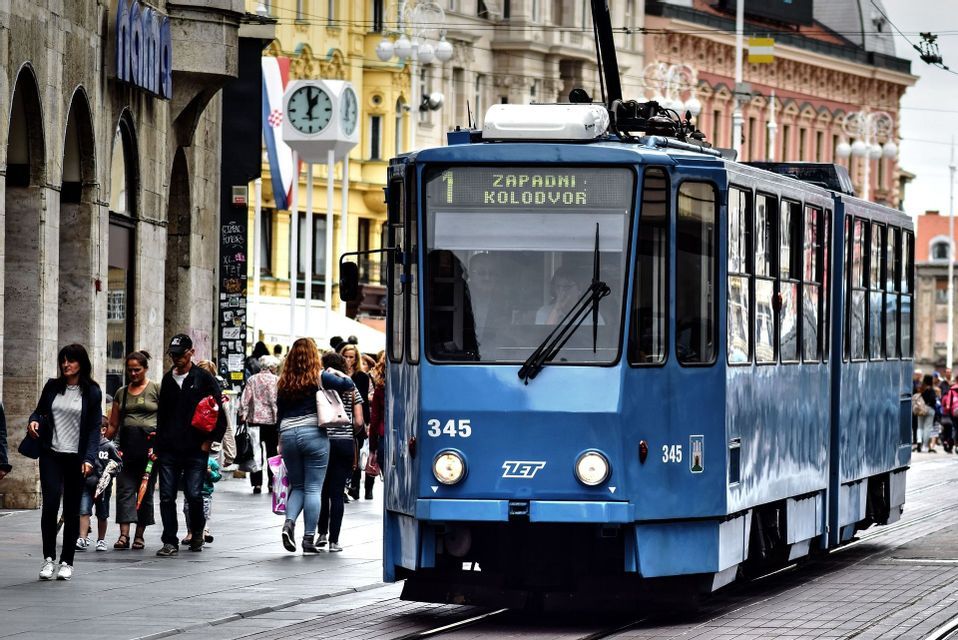 Un tram blu viaggia sui binari lungo una trafficata strada cittadina mentre le persone camminano sul marciapiede.