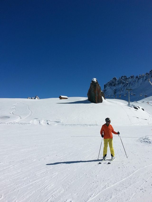 Una persona con una chaqueta naranja y pantalones amarillos está de pie sobre esquís en una ladera de montaña nevada bajo un cielo azul claro.