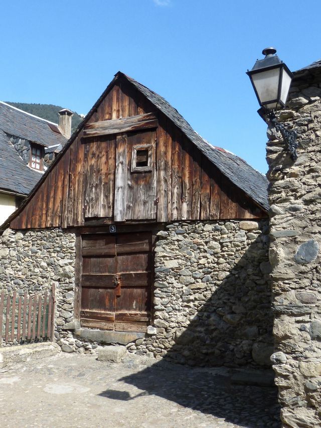 Un edificio rústico con base de piedra, fachada de madera desgastada y tejado de pizarra se alza bajo un cielo azul claro.