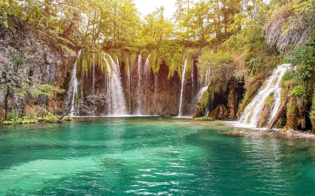 Diverse cascate scendono da una scogliera muschiosa in un lago limpido color turchese, circondato da una foresta rigogliosa e illuminata dal sole.