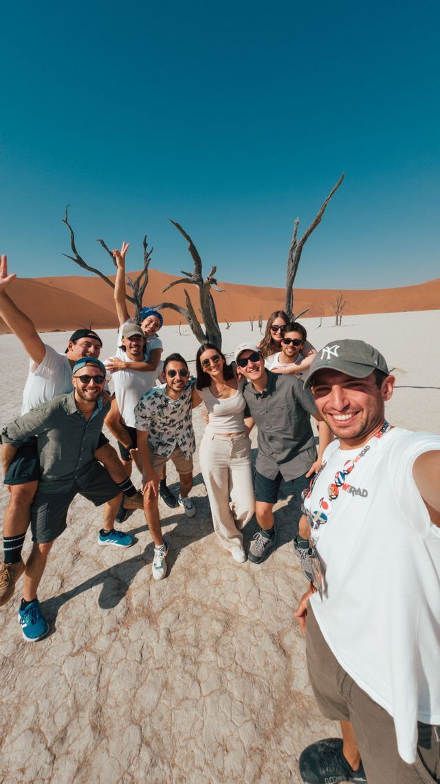 Eine WeRoad-Gruppe macht ein lächelndes Selfie in einer trockenen Wüste mit knorrigen Bäumen und orangefarbenen Sanddünen im Hintergrund.