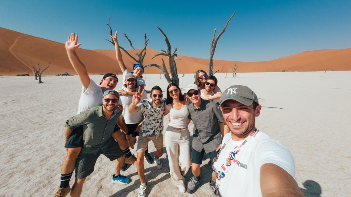 Eine WeRoad-Gruppenreise posiert für ein Selfie in einer Wüstenlandschaft mit toten Bäumen und großen orangefarbenen Sanddünen im Hintergrund.
