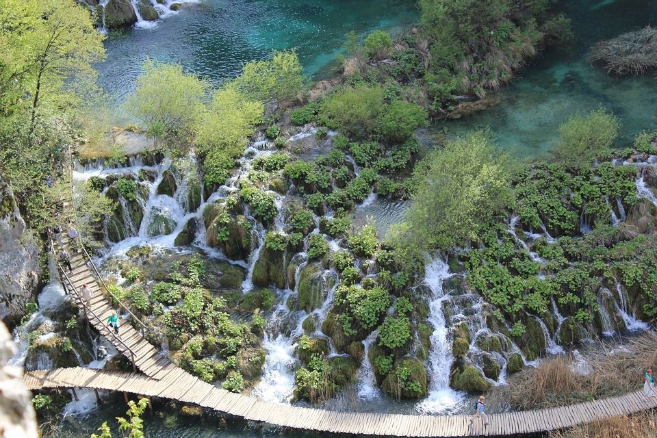 Una vista aerea di persone che camminano su una passerella di legno che si snoda tra cascate terrazzate e piscine turchesi.