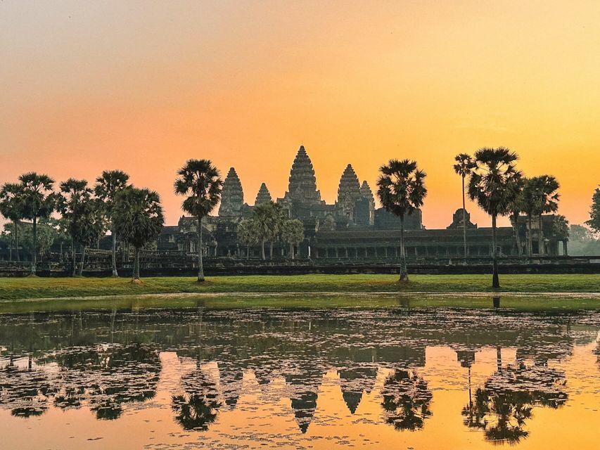 An ancient temple with tall spires and palm trees reflected in a calm body of water against an orange sky at sunrise.