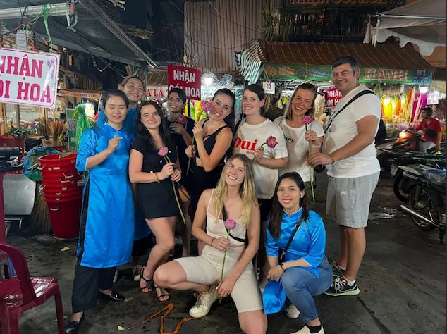 A WeRoad group trip poses together holding pink lotus flowers at a bustling outdoor night market.