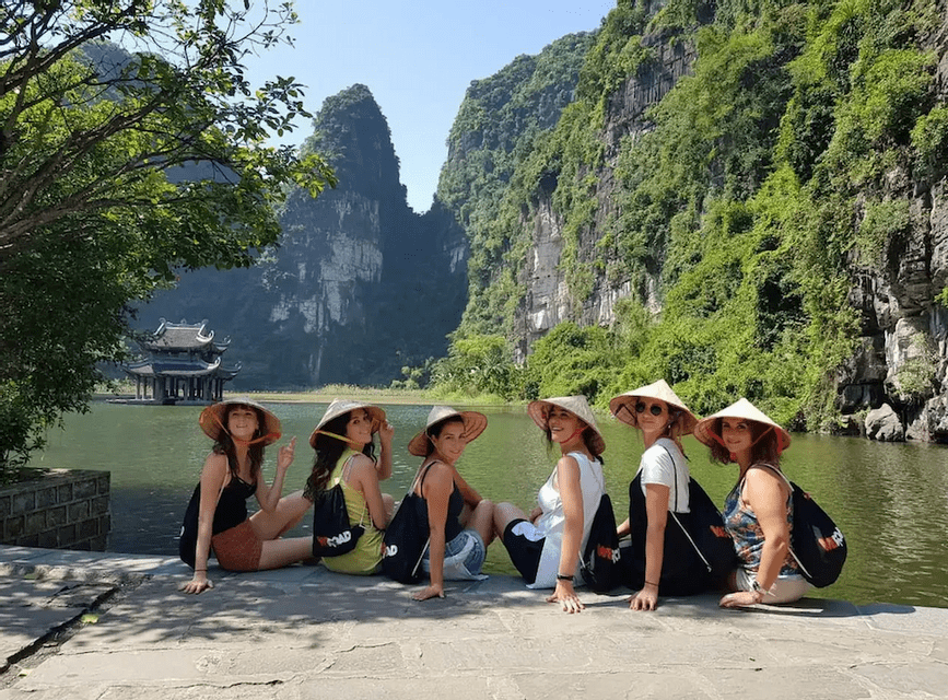 A WeRoad group trip of six women wearing conical hats, sitting by a river with green mountains in the background.