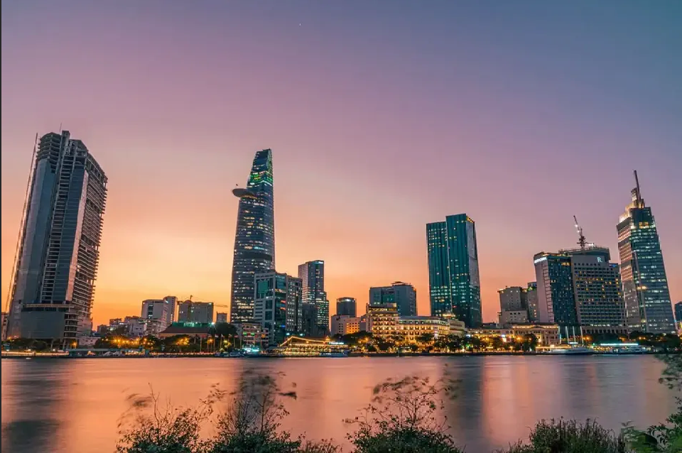 A modern city skyline with illuminated skyscrapers reflecting in a river against a colorful sunset sky.