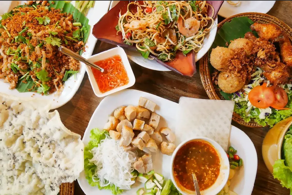 An overhead view of a table spread with various Southeast Asian dishes, including a noodle salad, fried foods, and dipping sauces.
