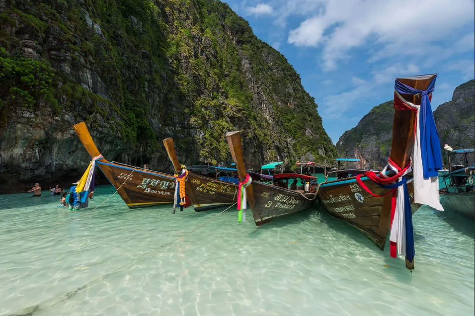Traditional long-tail boats with colorful ribbons float on clear turquoise water beside a large, green-covered cliff.
