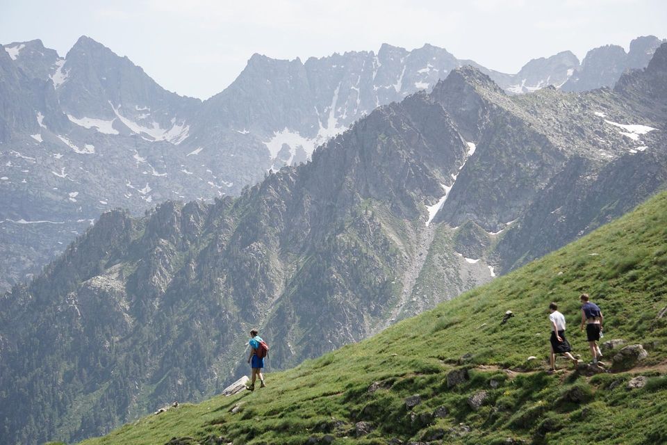 Un grupo de WeRoad de tres personas haciendo senderismo por una ladera de montaña empinada y herbosa, con picos escarpados y nevados de fondo.