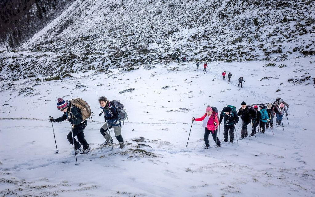 Un viaje en grupo de WeRoad haciendo senderismo en fila india por una ladera de montaña nevada, usando bastones de trekking y llevando mochilas.