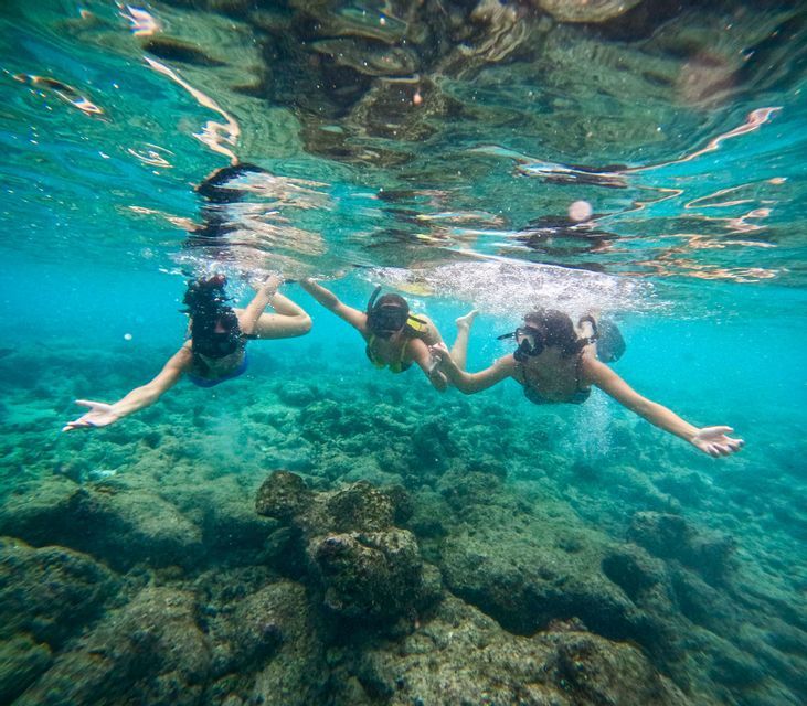 Drei Personen einer WeRoad-Gruppenreise schnorcheln gemeinsam, Hand in Hand in klarem türkisfarbenem Wasser über einem felsigen Meeresgrund.
