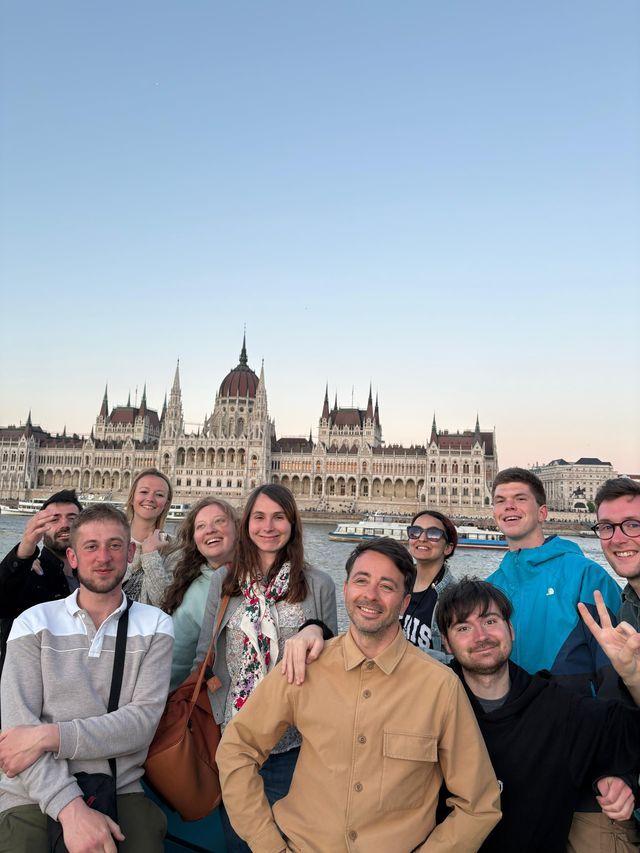 Un groupe WeRoad en voyage souriant pour une photo devant un grand bâtiment orné du parlement, de l'autre côté d'une rivière au crépuscule.