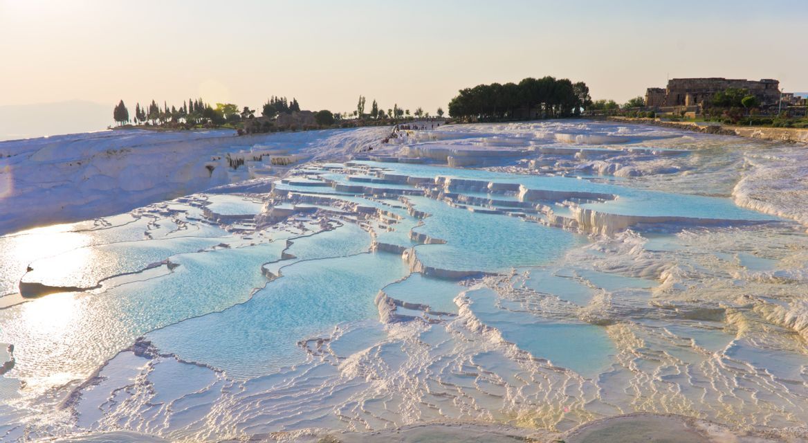 A scenic view of terraced thermal pools filled with turquoise water on a white mineral hill during a glowing sunset.