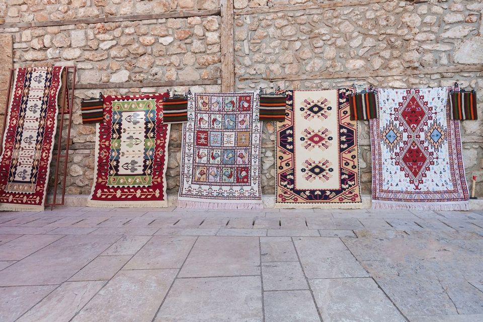 A collection of colorful, patterned rugs with intricate designs hanging on display against a rustic stone wall.