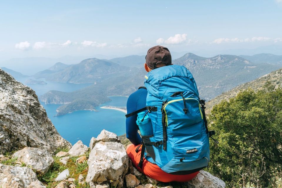 A hiker with a blue backpack sits on a rocky ledge, looking out over a coastal landscape of blue water and green, hilly islands.