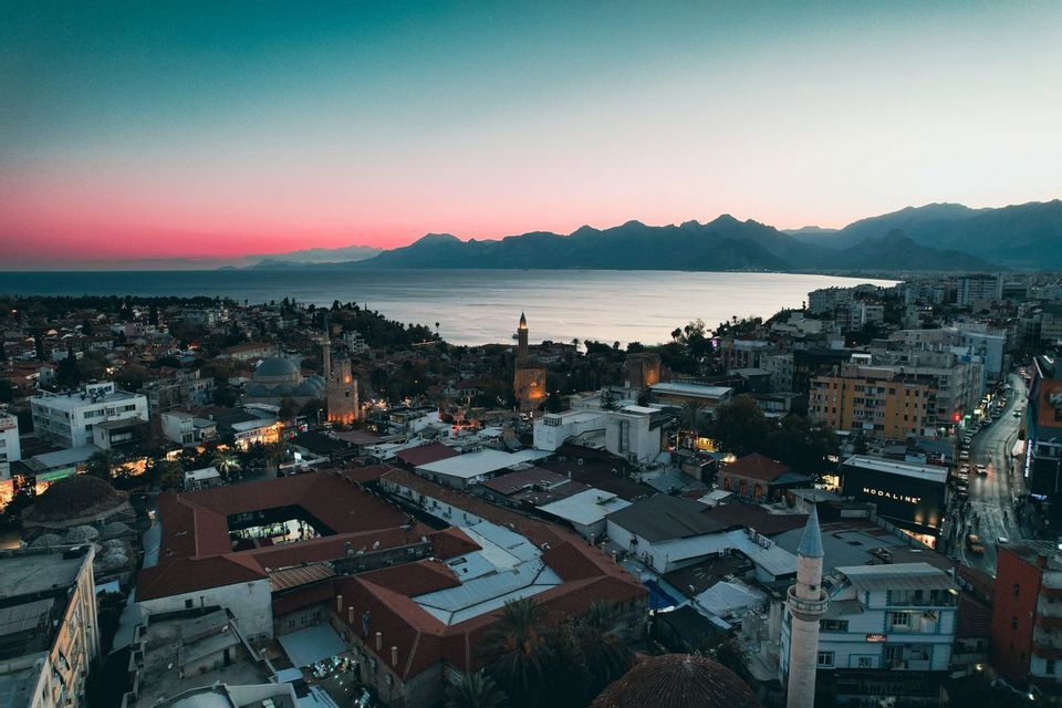 An aerial view of a seaside city with minarets and mountains under a pink and teal sky at dusk.