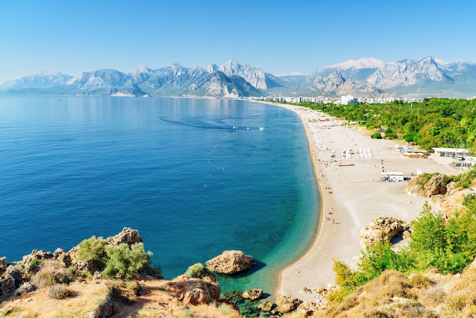 An aerial view of a long, curving beach with turquoise water, a coastal city, and a mountain range in the background under a clear sky.