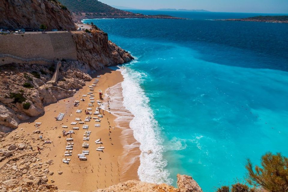 An aerial view of a sandy beach with white lounge chairs next to turquoise ocean waves at the bottom of a rocky cliff.