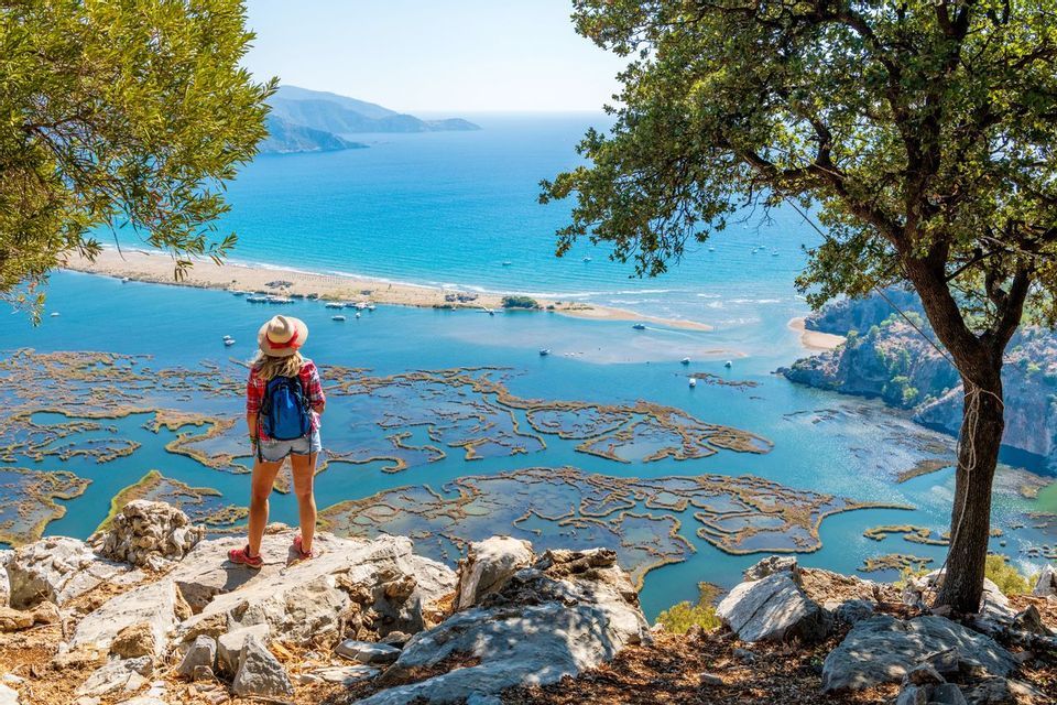 A person with a backpack and hat stands on a rocky clifftop, looking out over a coastal delta and a wide blue sea.