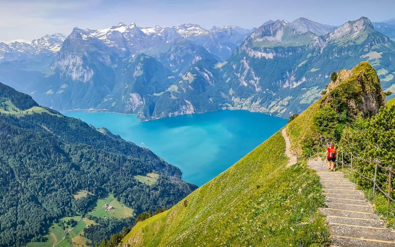 A hiker with a red backpack descends a steep mountain path overlooking a turquoise lake and distant snow-capped mountains.