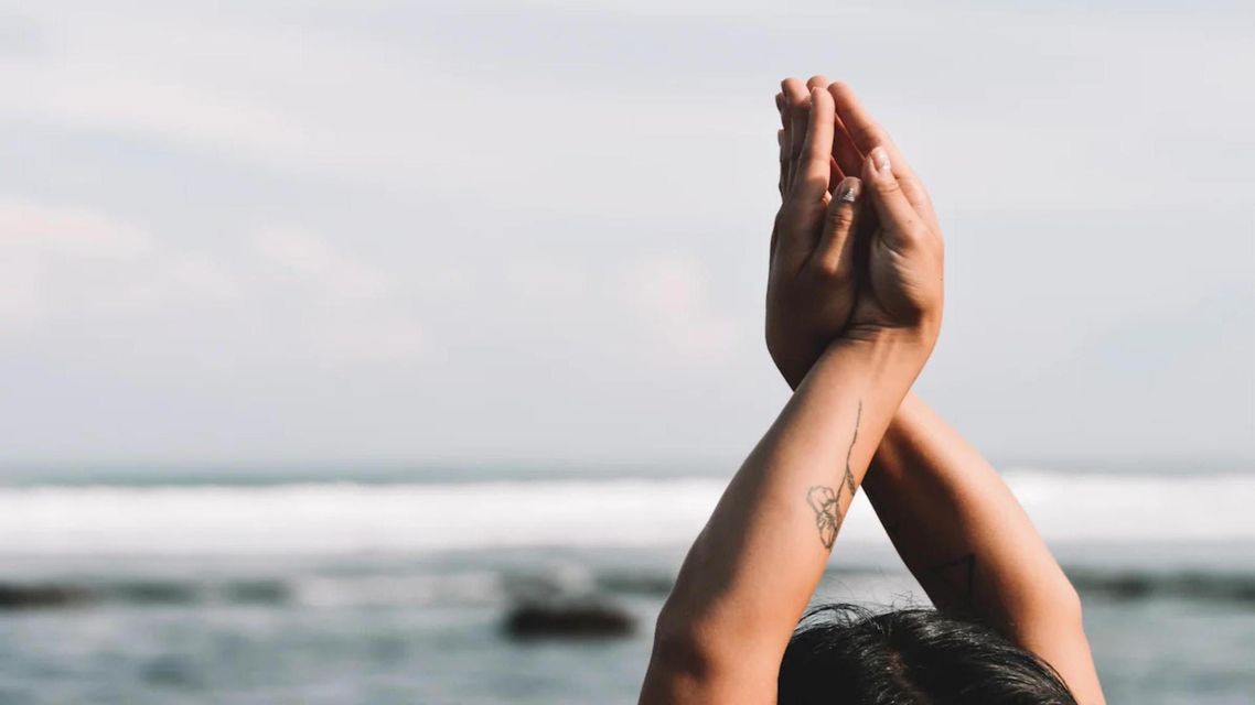 Un primo piano delle braccia di una persona con le mani giunte in una posa yoga davanti a un oceano sfocato.