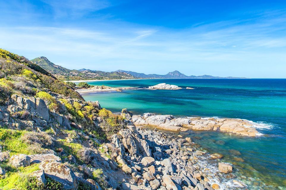 Una costa rocciosa con vegetazione verde e gialla si affaccia su un mare turchese, con una spiaggia sabbiosa e montagne visibili in lontananza.