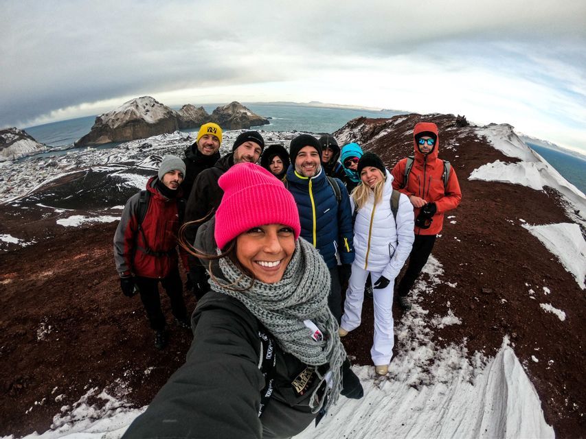 A woman in a pink beanie takes a selfie with her WeRoad group trip on a snowy, reddish-brown mountain overlooking the sea.