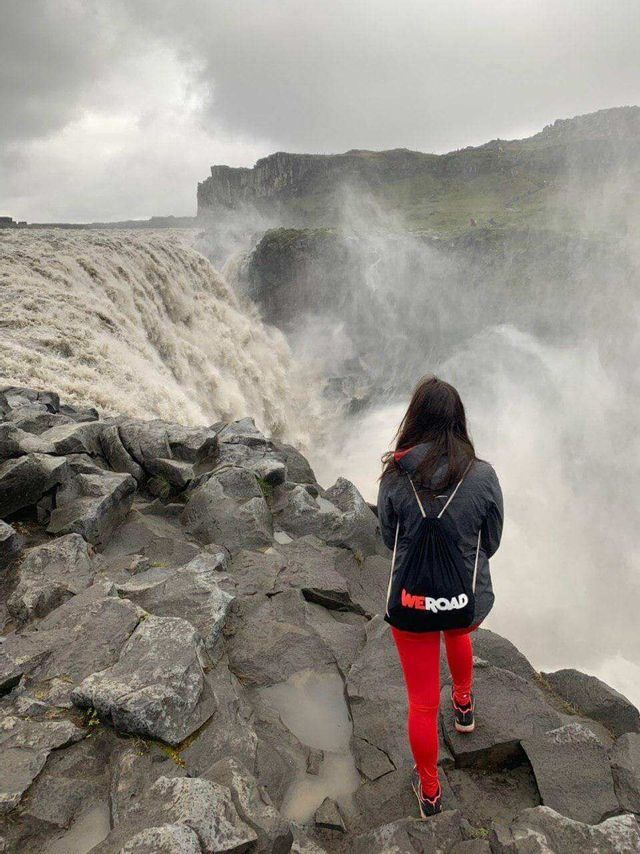 A woman seen from behind, wearing a WeRoad backpack, stands on rocks overlooking a massive, misty waterfall.
