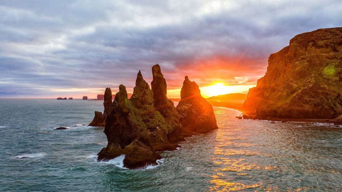 Jagged sea stacks covered in moss rise from the ocean as the sun sets, casting an orange glow on the water.