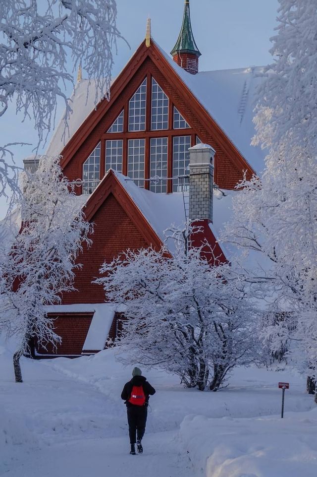 Eine Person mit rotem Rucksack wandert auf einem verschneiten Pfad zu einer großen, roten Holzkirche, umgeben von schneebedeckten Bäumen.