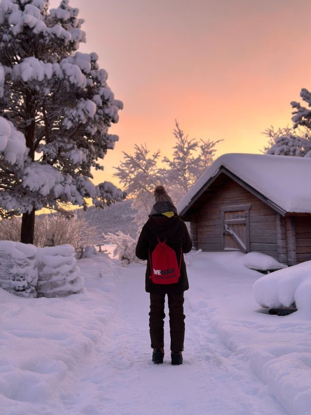 Eine Person mit rotem WeRoad-Rucksack steht bei Sonnenuntergang auf einem verschneiten Weg neben einer Holzhütte.