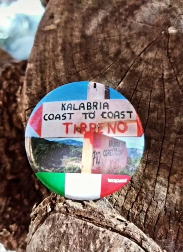 Un primo piano di una spilla WeRoad circolare per il sentiero 'Kalabria Coast to Coast', con un design a bandiera italiana, poggiata su un tronco di legno.