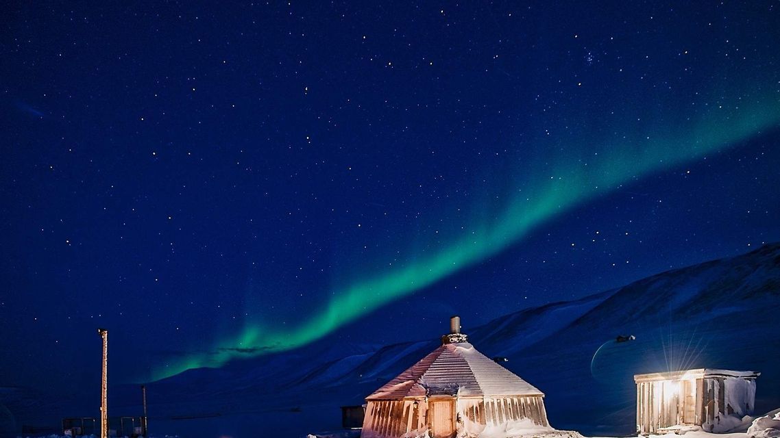 L'aurora boreale verde attraversa un cielo notturno stellato sopra baite e montagne innevate.