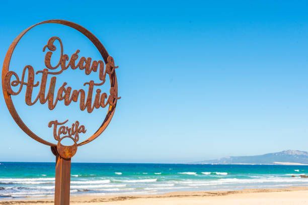 Un cartel metálico oxidado que dice 'Océano Atlántico Tarifa' se encuentra en una playa de arena con el océano turquesa de fondo.
