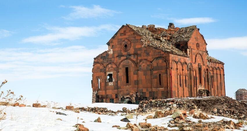 Una cattedrale diroccata in pietra rossa si erge su una collina innevata sotto un cielo azzurro splendente.