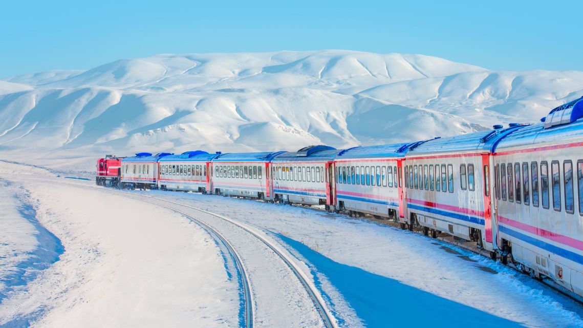 Un treno passeggeri viaggia su un binario curvo attraverso un paesaggio innevato con montagne sotto un cielo azzurro.