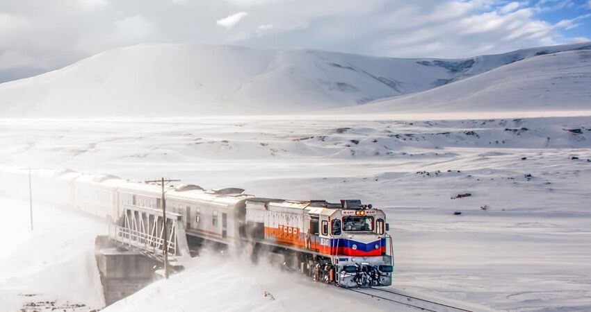 Un treno con una locomotiva rossa e blu attraversa un paesaggio innevato, sollevando neve mentre si muove.